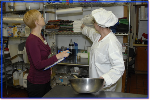 A Health inspector watching a cook and looking for wiping cloth storage in the prep area.